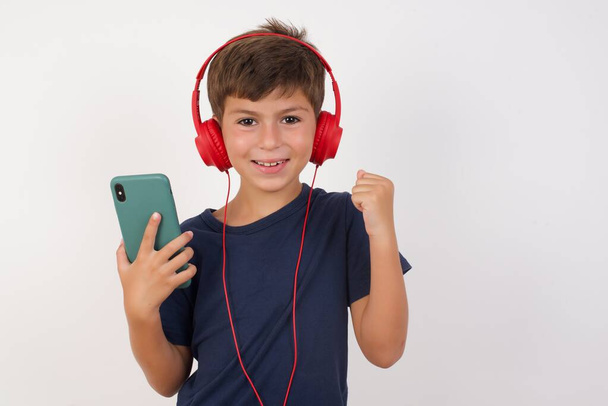 Positive Beautiful kid boy wearing casual t-shirt standing over isolated white background, holds modern cell phone connected to headphones, clenches fist from good emotions, exclaims with joy,  - Photo, Image
