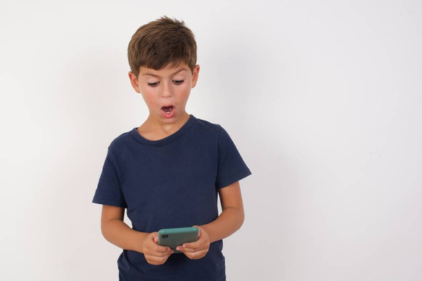 Portré fókuszált Gyönyörű gyerek fiú visel alkalmi póló áll elszigetelt fehér háttér, használja okostelefon olvasás szociális média hírek, vagy fontos e-mail  - Fotó, kép