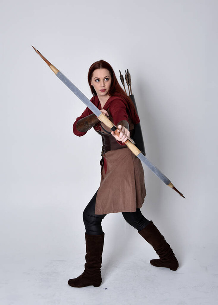 Портрет девушки с рыжими волосами в коричневом средневековом костюме лучника. Стоячая поза, держащая лук и стрелы, изолированные на сером фоне студии. - Фото, изображение