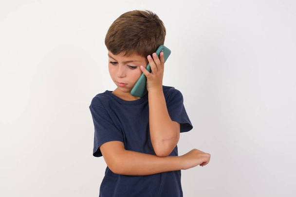Sad Beautiful kid boy wearing casual t-shirt standing over isolated white background, talking on smartphone. Communication concept. - Photo, Image