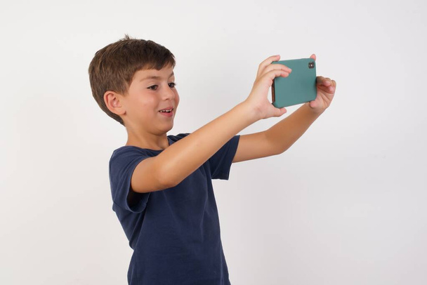 Κοντινό πλάνο πορτρέτο Όμορφο αγόρι φορώντας casual t-shirt στέκεται πάνω από απομονωμένο λευκό φόντο, βγάζοντας μια selfie για να το δημοσιεύσετε στα μέσα κοινωνικής δικτύωσης ή έχοντας μια βιντεοκλήση με φίλους ή κάποιο μέλος της οικογένειας. - Φωτογραφία, εικόνα