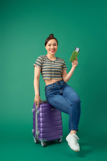 Attractive young Asian woman sitting on suitcase and holding passport, ticket flight over green background. - Foto, imagen