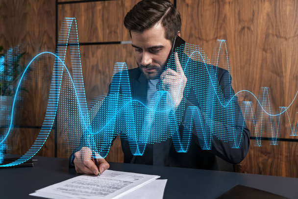 A man in office signing papers while talking phone and stock market graph and chart hologram. Double exposure. Formal wear. - Фото, изображение