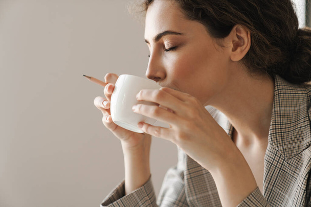 Photo closeup of pleased brunette woman drinking coffee while working in white office - Fotoğraf, Görsel