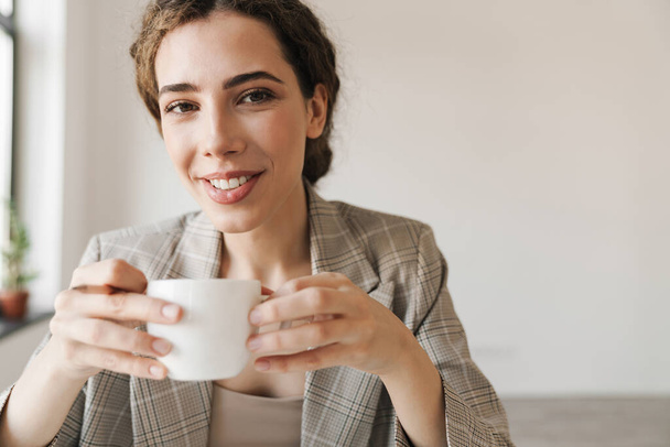 Photo closeup of smiling brunette woman drinking coffee while working in white office - Фото, изображение