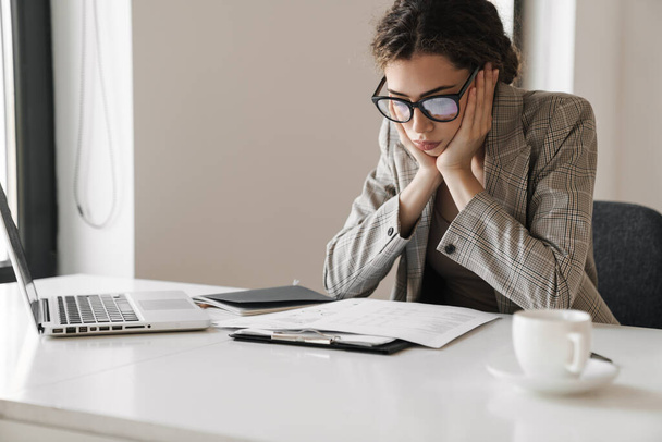 Photo of brooding brunette woman in eyeglasses working with papers and laptop in white office - Foto, afbeelding