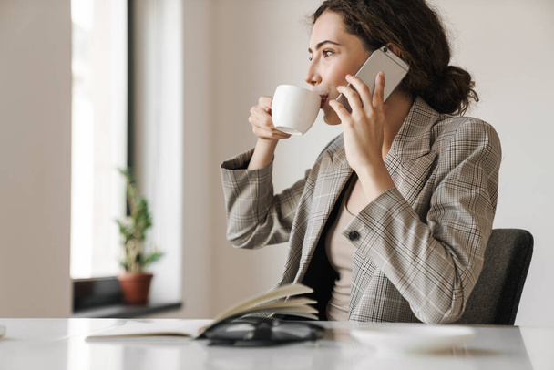 Photo of pleased brunette woman drinking coffee and talking cellphone while working in white office - Foto, imagen