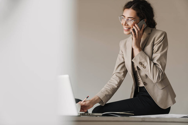 Photo of smiling brunette woman in eyeglasses talking on cellphone while working with laptop at office - Foto, afbeelding