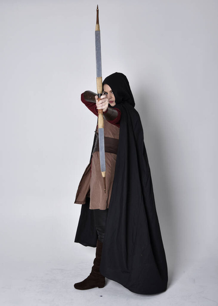 Πλήρες πορτρέτο του κοριτσιού με κόκκινα μαλλιά φορώντας μεσαιωνική στολή τοξότη με μαύρο μανδύα. Μόνιμη στάση με την πλάτη στην κάμερα κρατώντας ένα τόξο και βέλος, απομονώνονται σε ένα γκρι φόντο στούντιο. - Φωτογραφία, εικόνα
