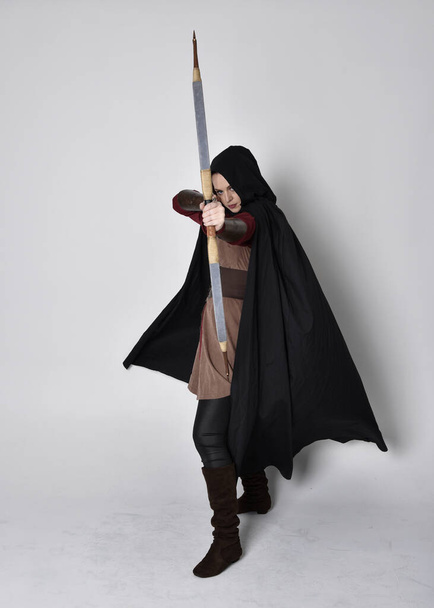 Full length portrait of girl with red hair wearing medieval archer costume with black cloak. Standing pose with back to the camera holding a bow and arrow,  isolated against a grey studio background. - Photo, Image