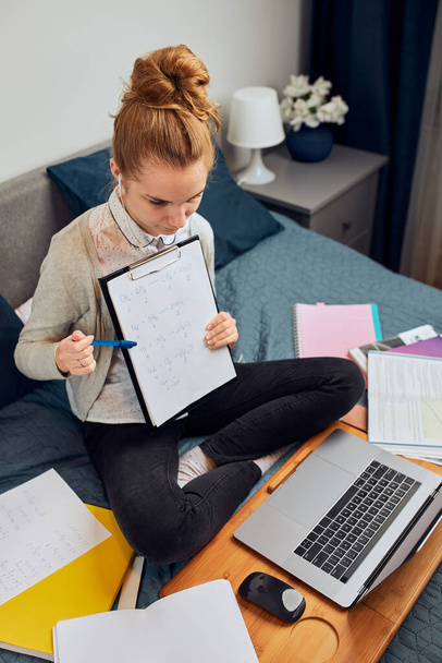 Young woman student having classes, learning online, watching lesson remotely, listening to professor, talking with classmates on video call from home during quarantine. Young girl using laptop, headphones, books, manuals sitting on bed - Foto, Imagen