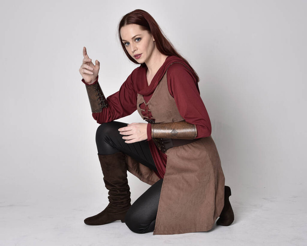 Retrato de cuerpo entero de chica de pelo rojo con traje de arquero medieval. Posada sentada, aislada sobre un fondo gris de estudio. - Foto, imagen