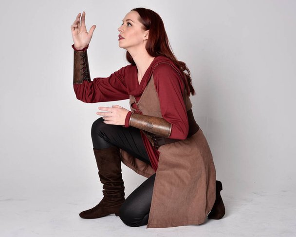 Retrato de cuerpo entero de chica de pelo rojo con traje de arquero medieval. Posada sentada, aislada sobre un fondo gris de estudio. - Foto, imagen