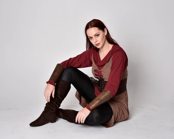 Портрет девушки с рыжими волосами в средневековом костюме лучника. Сидящая поза, изолированная на сером фоне студии. - Фото, изображение