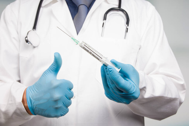 Detailní záběr ruky lékaře v lékařských rukavicích s nataženou injekční stříkačkou s vakcínou Covid-19 ukazující palce nahoru - Fotografie, Obrázek