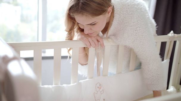 Happy mother near baby crib. Mom looking on sleeping child in cot - Zdjęcie, obraz