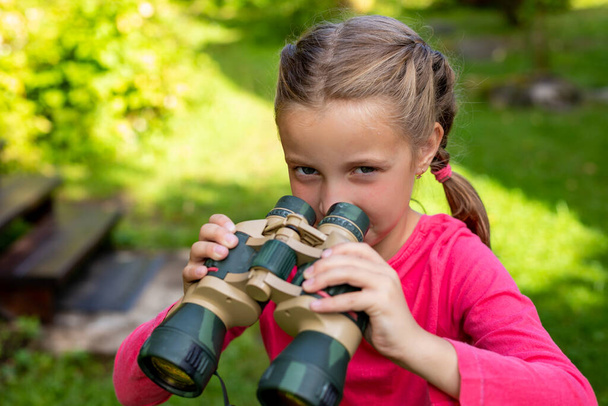 Blonde little girl looking through binoculars on nature background. baby hid in greenery and is sitting in ambush. Kids Adventure Concept. The child is watching the parents at a distance - Photo, Image