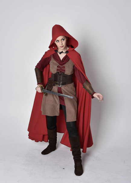 Retrato completo de niña vestida con traje medieval y capa roja. Posar de pie sosteniendo una espada, aislado sobre un fondo gris estudio. - Foto, Imagen