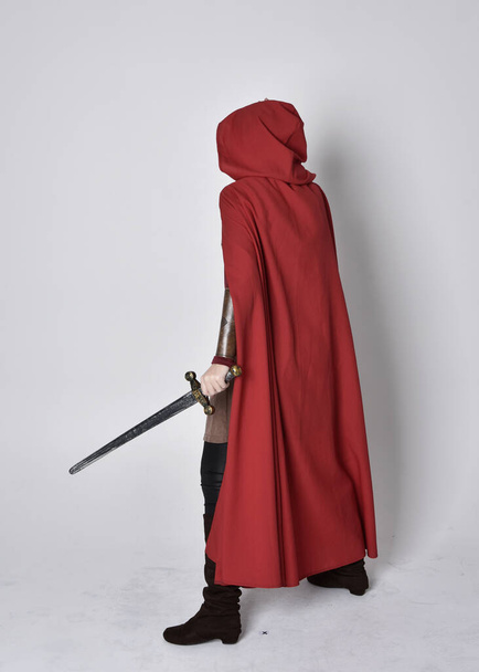 Dlouhý portrét dívky ve středověkém kostýmu a červeném plášti. Stojící póza s mečem, izolovaný na pozadí šedého studia. - Fotografie, Obrázek