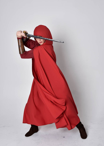 Portrait complet de fille portant un costume médiéval et un manteau rouge. pose debout tenant une épée, isolé sur un fond de studio gris. - Photo, image