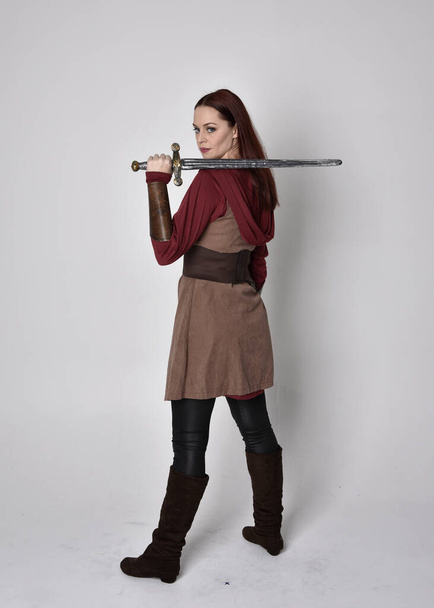 Full length portrait of girl wearing medieval costume. Standing pose holding a sword with back to the camera  isolated against a grey studio background. - Photo, Image