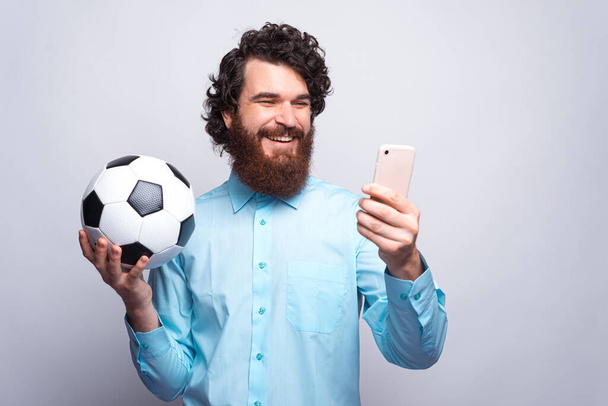 Πορτρέτο του χαμογελαστού άνδρα σε casual κρατώντας μπάλα ποδοσφαίρου και ψάχνει - Φωτογραφία, εικόνα