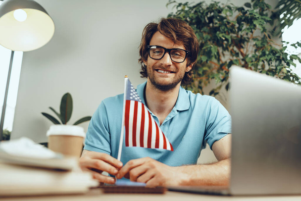 Young bearded businessman sitting at table with laptop and american flag - Fotoğraf, Görsel