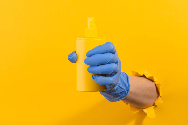 Hand in a latex glove giving a sunscreen spray through torn yellow wall - Фото, изображение