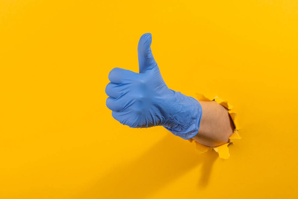 Hand in a latex glove showing a thumb up through torn yellow wall - Fotó, kép