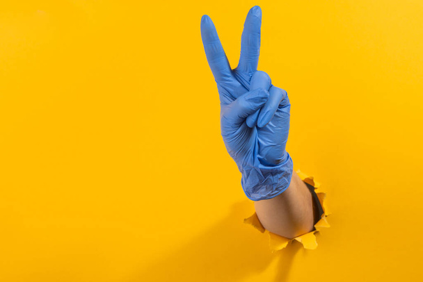 Hand in a latex glove showing a victory sign through torn yellow wall - Photo, image
