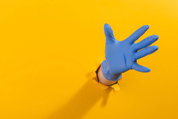 Hand in a latex glove grabbing something through torn yellow wall - Foto, Imagen
