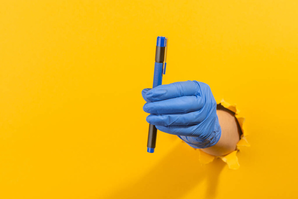 Hand in a latex glove holding a pen through torn yellow wall - Foto, Imagem