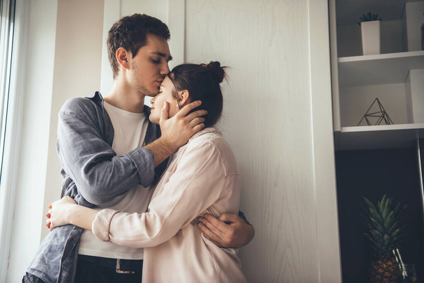 Linda pareja joven besándose en la cocina y abrazándose cerca de la ventana - Foto, imagen