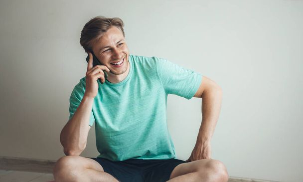 Usmívající se blondýnka muž má přestávku během lekce fitness doma a mluvit po telefonu - Fotografie, Obrázek