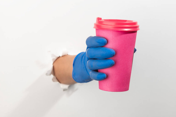 Hand in a latex glove giving a pink coffee cup through torn white wall - Foto, afbeelding