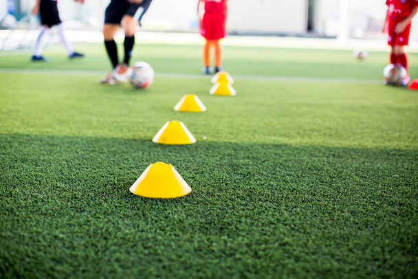 selektywne skupienie się na żółtym stożkowym markerze to sprzęt do treningu piłki nożnej na zielonej sztucznej trawie z rozmytym tłem treningowym dzieci. Materiał do szkolenia klasy akademii piłkarskiej. - Zdjęcie, obraz