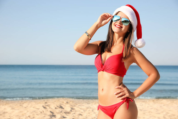 Young woman wearing Santa hat and bikini on beach, space for text. Christmas vacation - Zdjęcie, obraz