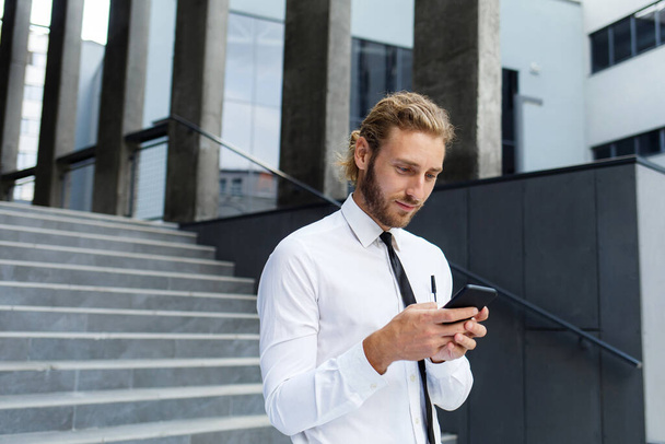 Portrait of a successful young businessman. A curly-haired man in a white shirt with a telephone against the background of a modern business center - Foto, afbeelding