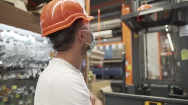 Muž pracující ve skladu, inventář obchodu, a provozuje vysokozdvižný vozík. Stavební dělník v supermarketu v oranžové helmě, brýlích a masce během coronoviris a covid 19 pandemie - Záběry, video
