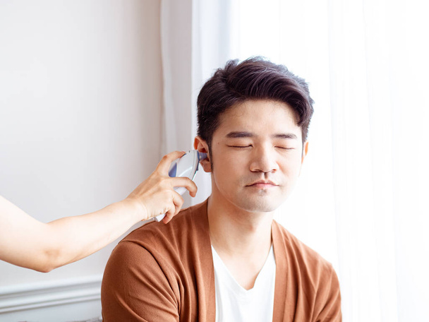 Jeune homme asiatique portant un masque chirurgical et utilisant un thermomètre auriculaire - Photo, image