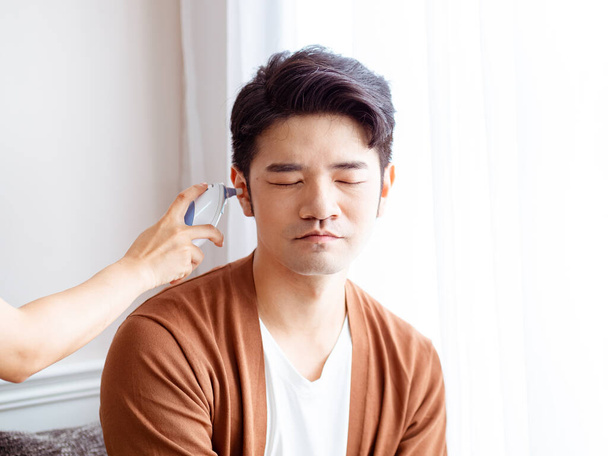 Jeune homme asiatique portant un masque chirurgical et utilisant un thermomètre auriculaire - Photo, image