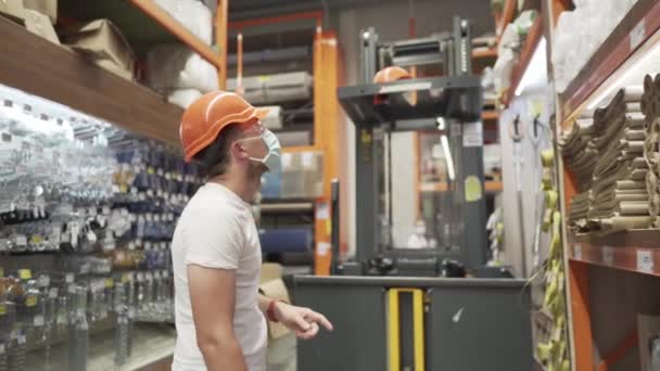 Trabalhador industrial caucasiano em chapéu duro laranja, óculos de segurança e máscara médica na loja de ferragens contra o fundo de um elevador de carga de carga. masculino armazém trabalhador máscara coronavírus, covid 19 - Filmagem, Vídeo