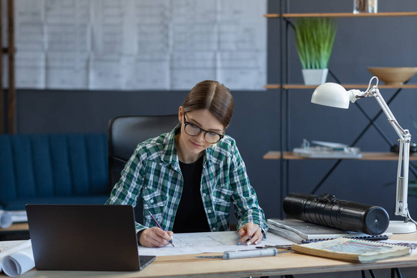 Female interior designer working in office with laptop. Architect thinks over architectural plan, searching new ideas for construction project. Woman sitting at workplace. Business portrait concept - Φωτογραφία, εικόνα