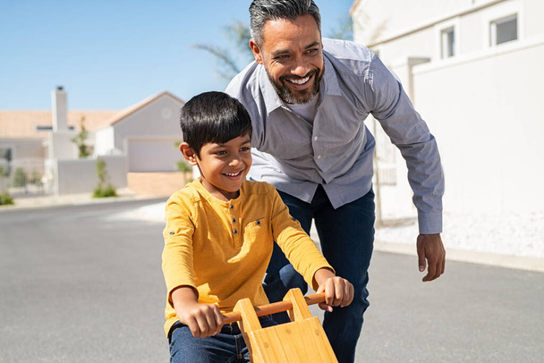 Cheerful middle eastern father helping excited son to ride wooden balance cycle on street. Happy boy enjoying riding bycicle with his dad. Dad teaching his indian son to ride bicycle outdoor. - Foto, afbeelding