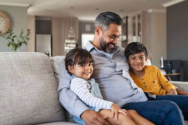 Happy daughter and smiling son relaxing with father on couch at home. Lovely man embracing boy and girl sitting on sofa. Cheerful indian family with two children relaxing at cozy home with copy space. - Foto, Imagem