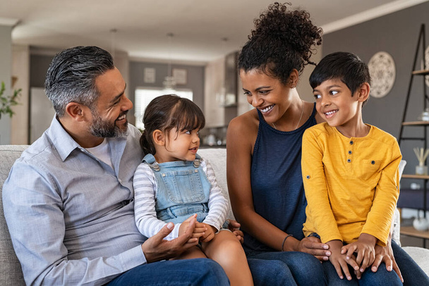 Happy latin family sitting on sofa and laughing together. Cheerful middle eastern parents playing with their daughter and son at home. Indian father sitting with daughter while african mother and brother smiling. - Foto, Imagem