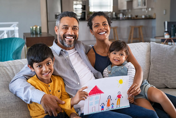 Proud parents showing family painting of son and daughter sitting on sofa at home. Smiling african mother and middle eastern father with two children looking at camera. Little boy and cute girl with parents showing handmade painting of a happy united - Photo, Image