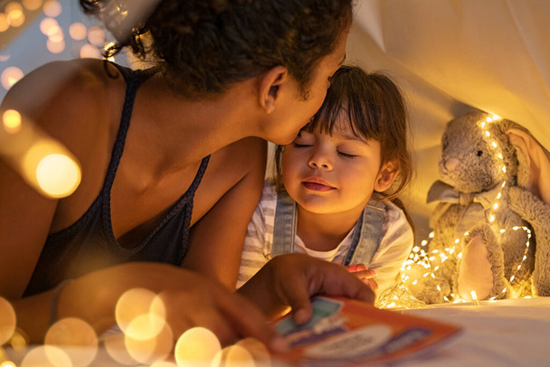 African american mother kissing her daughter on forehead while lying on bed in illuminated tent. Close up of mom kissing little cute girl with closed eyes holding story book in tent. Mother putting daughter to sleep in cozy hut after reading a fairy  - Foto, imagen