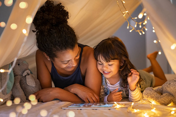African mother and cute smiling girl using digital tablet while lying in illuminted tent in kid bedroom. Cheerful ethnic woman and lovely daughter on video call under a cozy hut. Lovely little girl with mom watching cartoon on digital tablet in bedro - Фото, изображение