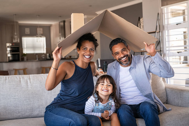 Portrait of smiling family sitting on couch holding cardboard roof and looking at camera. African and indian parents with funny daughter holding cardboard roof over heads while sitting on sofa in their new home. Happy mother and father with daughter  - Zdjęcie, obraz